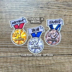 May include: Three circular stickers shaped like medals with ribbons. The left medal is gold with #1 APPRENTICE and the name Matthew. The center is silver with #2 APPRENTICE and the name Jacob. The right is bronze with #2 APPRENTICE and the name Seaweed.