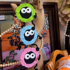 May include: Three handmade crochet monster ornaments in green, blue, and pink. Each has a black face with white eyes and black legs. The ornaments are hanging from a wooden hand, with a pearl necklace and framed artwork in the background.