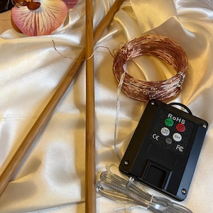 May include: Two long, smooth, light brown wooden knitting needles positioned diagonally next to a coiled strand of thin copper wire fairy lights and a black rectangular battery pack with buttons labeled RoHS, MODE, ON/OFF, TIME OFF, and 6H.