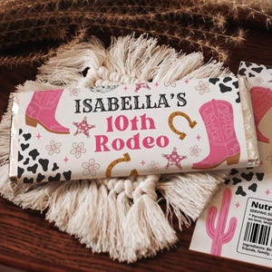 Può includere: Confezione di barretta di caramelle a tema rodeo. Presenta stivali da cowboy rosa, stelle, ferri di cavallo e il testo "ISABELLA'S 10th Rodeo". Lo sfondo include una stampa di mucca ed elementi floreali. La barretta poggia su un sottobicchiere in macramè.