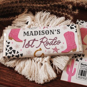May include: A chocolate bar wrapper with the text "MADISON'S 1st Rodeo" in pink and brown. The wrapper features pink cowboy boots, flowers, a horseshoe, and a star. Black and white cow spots are also present. The wrapper is on a macrame coaster.