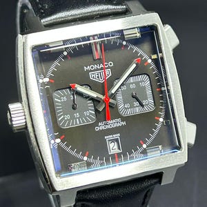 May include: A square-cased Monaco Heuer automatic chronograph watch with a black dial, two sub-dials, a red central second hand, and a date window at the six o'clock position. The watch features a black leather strap and silver-tone metal casing.