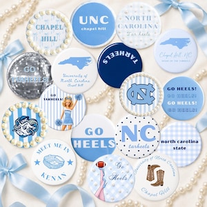 Può includere: Una collezione di bottoni rotondi con vari disegni e testo, principalmente in tonalità di blu e bianco. Alcuni bottoni presentano il logo UNC, le parole "Chapel Hill" e "Tar Heels". Altri bottoni hanno una grafica di cheerleader e la scritta "Go Heels!"