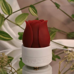 Pode incluir: Um objeto em forma de rosa vermelho-escuro com pétalas lisas e arredondadas assenta sobre uma base cilíndrica branca. A base apresenta um padrão de videira subtilmente em relevo na sua circunferência e um pequeno componente metálico.