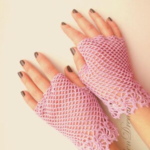 May include: A pair of pink crocheted fingerless gloves with a delicate lace trim. The gloves are made with a mesh pattern and have a light pink color.