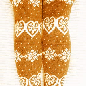 May include: Knee-high brown knit socks with white snowflake and heart patterns. The socks have a ribbed cuff and are visible from the knees down. The text "WoolenDream" is printed on the socks.
