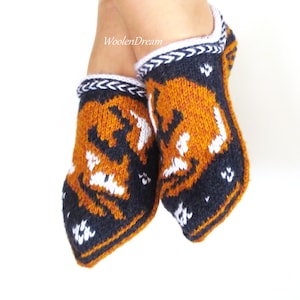 May include: A pair of knitted slippers with a fox design. The slippers are primarily orange and navy blue, with white accents. The top edge has a white and navy blue braided trim. The text "WoolenDream" is visible.