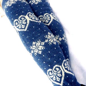 May include: A pair of blue knit leg warmers with white snowflake and heart patterns. The leg warmers are made of a soft, textured material and are designed to be worn over the lower legs.