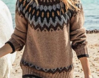 May include: A brown sweater with a geometric pattern in black, gray, and tan. The sweater has a high neckline and long sleeves with black trim. The background shows a beach scene with water and sand.