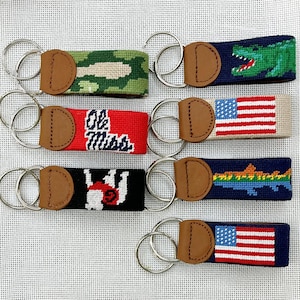 May include: A collection of seven rectangular needlepoint keychains featuring various designs including camouflage, a green alligator, the text Ole Miss, two American flags, a white bear, and a colorful fish, each with a brown leather tab and metal ring.