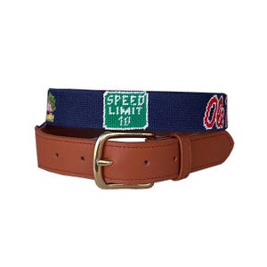 May include: A brown leather belt with a brass buckle. The navy blue fabric belt features a "Speed Limit 18" sign and the letters "Ole Miss". A stylish accessory for any outfit.