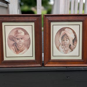 Puede incluir: Dos marcos cuadrados de madera con acabado marrón oscuro, cada uno con un retrato en tono sepia. El retrato de la izquierda muestra a un niño con sombrero de ala ancha y pañuelo. El de la derecha muestra a un niño con una capota atada.