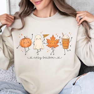 Könnte beinhalten: Ein cremefarbenes Sweatshirt mit Rundhalsausschnitt und einem Grafikdruck von vier Zeichentrickfiguren: einem orangefarbenen Kürbis, einem weißen Geist mit einem Wimpel, einem orangefarbenen Ahornblatt und einem braunen Kaffeebecher. Darunter steht der Text "cozy season".