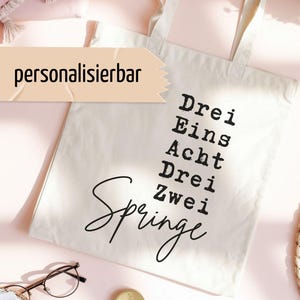 Könnte beinhalten: Eine cremefarbene Stofftasche aus Segeltuch mit schwarzem Aufdruck, der die Wörter Drei, Eins, Acht, Drei, Zwei und das Wort Springe in Schreibschrift zeigt. Ein rechteckiges Etikett mit dem Wort personalisierbar ist oben links angebracht.