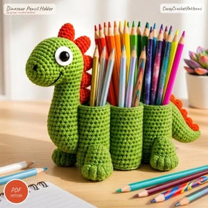 May include: A green crocheted dinosaur pencil holder with a red spine, large white and black eyes, and three cylindrical compartments filled with various colored pencils. The text Dinosaur Pencil Holder and CozzyCrochetPatterns is visible.