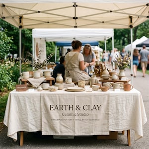 May include: A display table covered with a cream-colored cloth featuring the text EARTH & CLAY Ceramic Studio, Handmade Pottery, Small Batch Ceramics. The table holds various ceramic mugs, bowls, and vases in neutral tones under a white canopy.