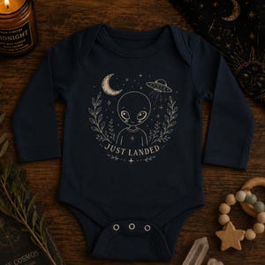 May include: A dark navy blue long-sleeve baby bodysuit featuring a cream-colored graphic of an alien, a crescent moon, a flying saucer, and leafy branches. The text "JUST LANDED" is printed below the alien figure, surrounded by small stars.