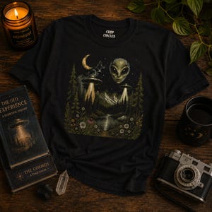 Könnte beinhalten: Ein schwarzes T-Shirt mit einem Grafikdruck eines Alien-Kopfes, zwei fliegenden Untertassen, die Lichtstrahlen über einen Wald aussenden, einer Mondsichel und einem See. Das T-Shirt hat einen Rundhalsausschnitt und den Text Crop Circles.