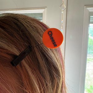 May include: A hair clip featuring a small, round, bright orange disc attached to a thin metal wire. The word "Cheese" is printed in bold, black, sans-serif capital letters across the center of the orange disc. The clip is a standard black metal.