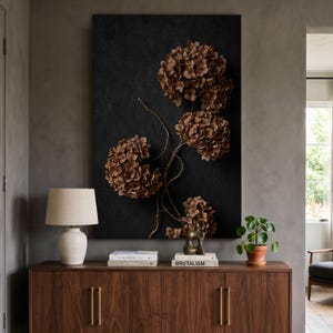 May include: A large rectangular wall art piece featuring dried brown hydrangea flowers on a textured black background. The artwork is displayed above a wooden sideboard with a white lamp, stacked books, a small sculpture, and a potted plant.