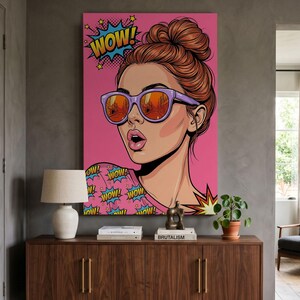 May include: A pop art style canvas print featuring a stylised illustration of a person with a high bun hairstyle wearing purple sunglasses with orange lenses. The background is bright pink with the word WOW! in a blue comic book speech bubble.