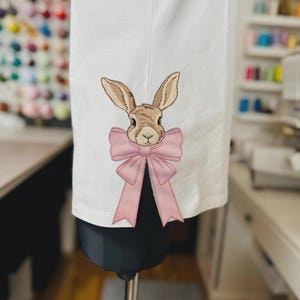 May include: A white fabric item featuring an embroidered brown rabbit head centered above a large, light pink bow. The rabbit has detailed fur texture and long ears, while the bow has defined folds and long, hanging ribbon tails.