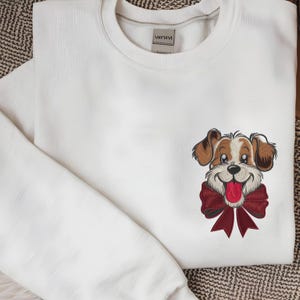 May include: A white crewneck sweatshirt featuring an embroidered graphic on the chest of a brown and white dog with a large dark red bow around its neck. The dog has a happy expression with its tongue out. A brand label is visible at the neckline.
