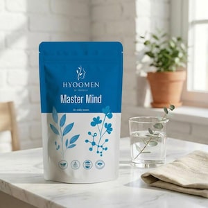 Op de afbeelding: Een blauw-wit zakje met de tekst "HYOOMEN Master Mind" en bloemmotieven. Een glas water met een takje groen en een beige doek liggen naast het zakje. Een plant in een terracotta pot staat op de achtergrond.