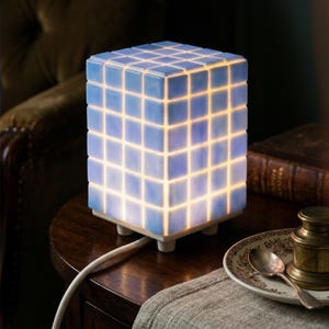 May include: A cube-shaped table lamp with a grid pattern of light blue squares. The lamp is illuminated from within, creating a soft glow. It sits on a white base with small feet and is connected to a white cord. The lamp is on a dark wooden table.