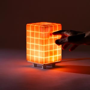 Puede incluir: Lámpara cúbica con un patrón de cuadrícula, que emite un brillo naranja cálido. La lámpara se asienta sobre una base blanca y está conectada a un cable de alimentación. Una mano se extiende hacia la superficie iluminada.