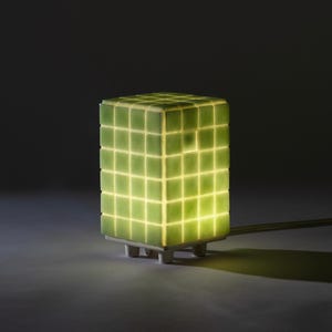 May include: A green, cube-shaped lamp with a grid pattern. The lamp is illuminated from within, creating a soft glow. It has a white base with four small feet and a green power cord.