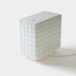 Könnte beinhalten: Eine weiße, rechteckige Lampe mit Mosaikfliesen-Design. Die Lampe besteht aus kleinen, quadratischen Fliesen, die eine strukturierte Oberfläche bilden. Sie hat ein weißes Kabel und steht auf einer weißen Oberfläche.