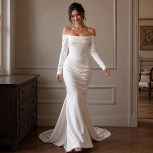 May include: A floor-length off-the-shoulder white gown with long sleeves, a structured corset-style bodice, and a draped skirt that flows into a train. The dress features a smooth, solid fabric finish and a fitted silhouette that flares slightly.