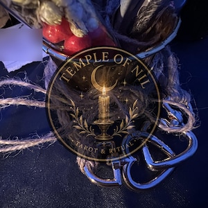 May include: A glass container wrapped in coarse brown twine, containing dried botanical elements and red spheres. Attached to the base are two polished silver-toned metal heart-shaped carabiners. A circular logo features a candle and the text TEMPLE OF NIL TAROT & RITUAL.