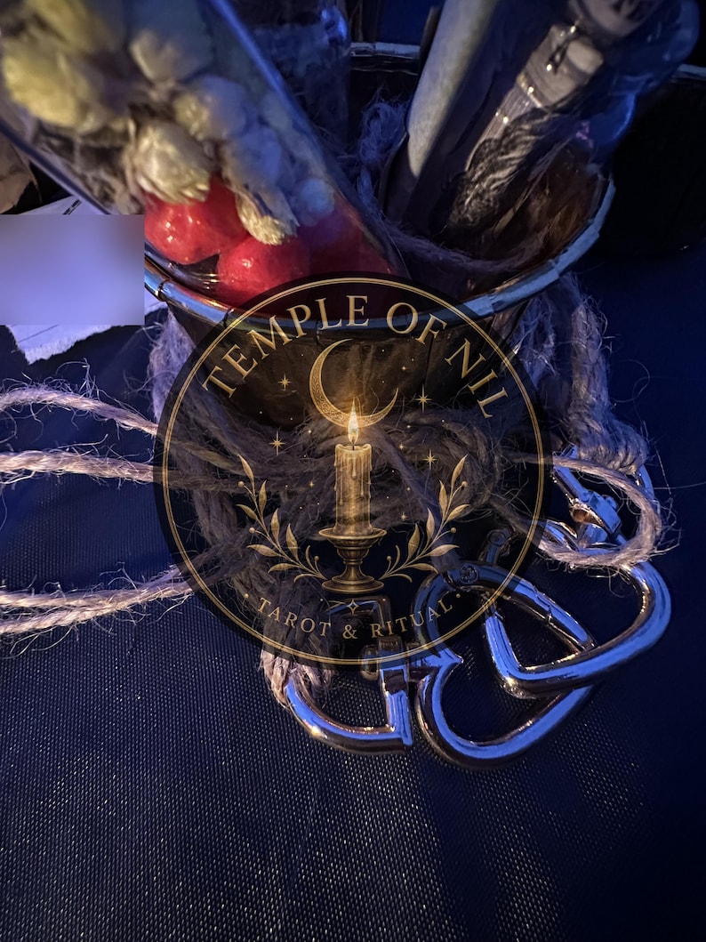 May include: A glass container wrapped in coarse brown twine, containing dried botanical elements and red spheres. Attached to the base are two polished silver-toned metal heart-shaped carabiners. A circular logo features a candle and the text TEMPLE OF NIL TAROT & RITUAL.