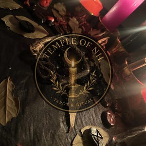 May include: A dark surface featuring scattered dried leaves, small black rune stones, and a glass vial filled with red items. Two lit candles, one orange and one pink, provide illumination. A circular logo reads Temple of Nil Tarot & Ritual.