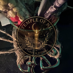 May include: A glass container filled with dried flowers and a rolled paper note, wrapped in natural jute twine. The container features a circular logo with the text TEMPLE OF NIL TAROT & RITUAL, depicting a crescent moon and a lit candle on a stand.