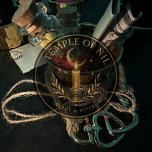 May include: A collection of items including a glass bottle with a cork stopper, a purple candle in a gold-toned holder, and a rolled paper note. A metallic heart-shaped clasp is attached to a piece of twine. The text Temple of Nil Tarot & Ritual is visible.