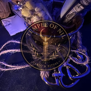 May include: A glass bottle filled with small white dried flowers, a dark metal container, and a silver-toned metal clasp attached to a piece of natural jute twine. The image features a circular logo with the text Temple of Nil Tarot & Ritual.