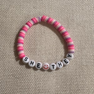 May include: A circular bracelet made of small, flat, round clay beads in shades of pink, light pink, and lavender. The bracelet features white letter beads spelling out the words SHE THEY with a small red smiley face bead between the words.