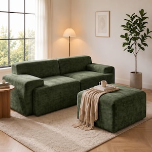 May include: A dark green velvet sofa and a matching square ottoman. The sofa features a low-profile design with wide armrests. A beige fringed throw blanket is draped over the ottoman, which also holds a white ceramic mug and a book.