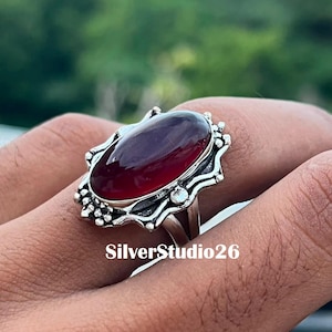 Op de afbeelding: Een zilveren ring met een grote, ovale, dieprode cabochon edelsteen in een decoratieve, geschulpte metalen zetting met kleine kraalaccenten. De tekst SilverStudio26 is zichtbaar aan de onderkant van de ringzetting.
