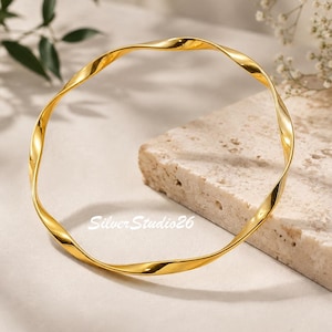 Può includere: Un bracciale rigido color oro lucido caratterizzato da un design continuo ritorto e ondulato. Il cerchio in metallo presenta una superficie liscia e riflettente, con il testo SilverStudio26 impresso in un piccolo carattere bianco senza grazie in basso.