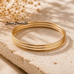 Puede incluir: Un conjunto de tres brazaletes de metal fino, liso y de color dorado. La joyería presenta un diseño circular simple con un acabado pulido. El texto SilverStudio26 es visible en letras negras cerca de los brazaletes sobre la superficie.