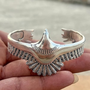 May include: A silver-toned cuff bracelet featuring a central eagle design with outstretched wings. The bird's plumage is depicted with detailed, etched line patterns across the wings and tail feathers, while the head remains smooth.
