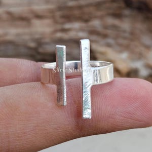 May include: A silver ring featuring an open band design with two vertical, rectangular bars of varying heights extending upwards from the shank. The metal surface has a textured, brushed finish and the text SilverStudio is visible on the band.