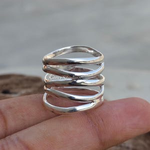 May include: A wide sterling silver ring featuring a multi-band design with five distinct, polished, and interconnected silver loops that wrap around the finger. The ring has a smooth, reflective surface and includes the text Silver Studio2c.