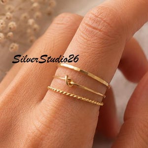 Può includere: Un set di tre sottili anelli d'oro indossati su un dito. L'anello superiore è una fascia liscia, l'anello centrale presenta un piccolo dettaglio a nodo e l'anello inferiore ha una trama a corda intrecciata. Il testo SilverStudio26 è visibile.