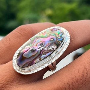 Puede incluir: Un anillo ovalado con una gran incrustación de concha de abulón iridiscente con tonos arremolinados de morado, verde y azul. La concha está engastada en un marco de plata texturizada con un borde decorativo. El texto SilverStudio26 es visible.