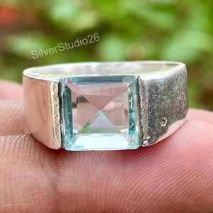May include: A silver ring featuring a square-cut, light blue gemstone set in the center. The band has a smooth, polished finish on the sides. The text SilverStudio26 is visible above the ring. The design is simple and focuses on the stone.
